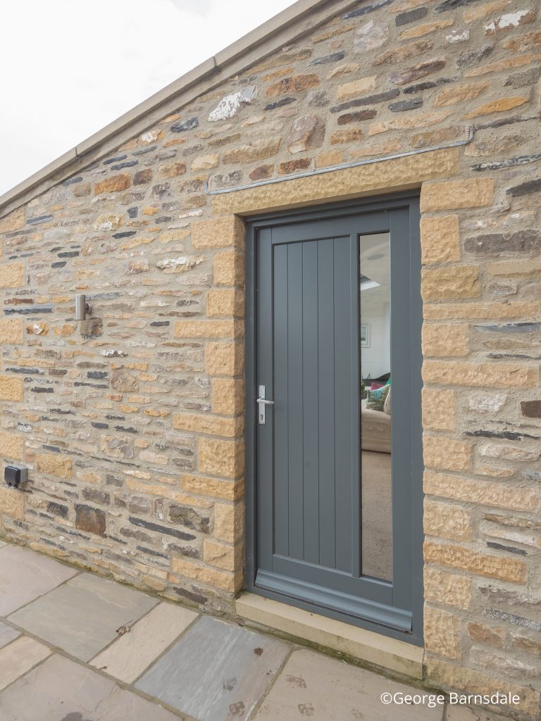 Grey timber George Barnsdale door with vertical panels and side glazing, set in a rustic stone wall.