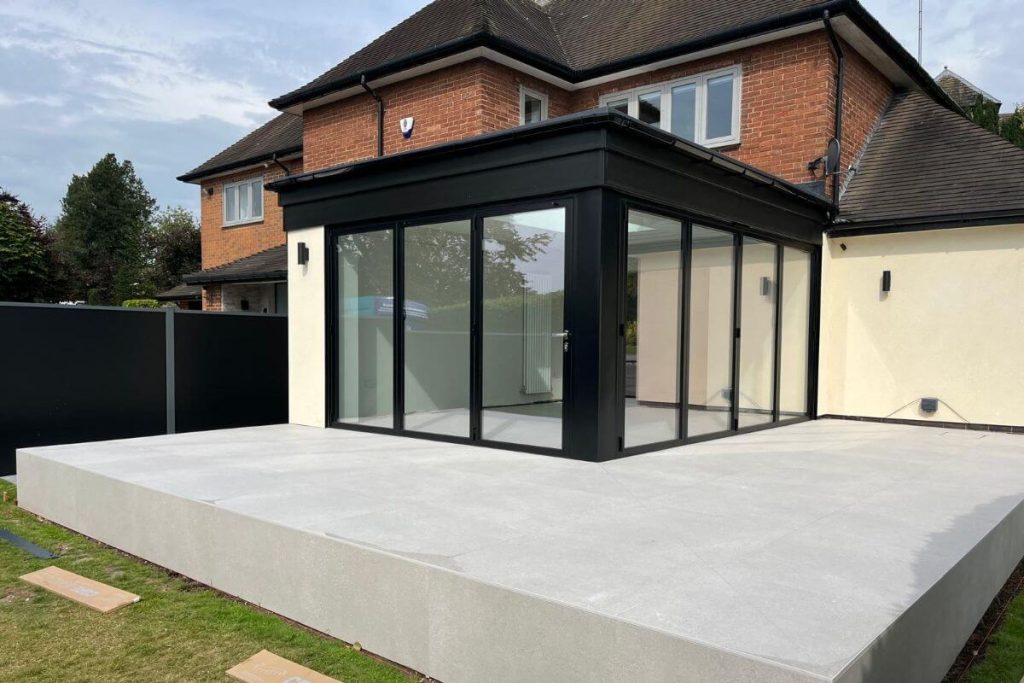 Angled view of a modern orangery with slimline black bifold doors and a smooth, elevated tiled patio extending into the garden.