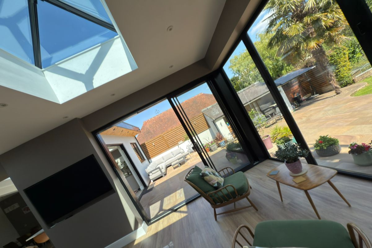 Interior view of a garden room with sliding glass doors and a roof lantern, opening out to a spacious patio. The room features modern seating, a small coffee table, and views of the landscaped outdoor area.