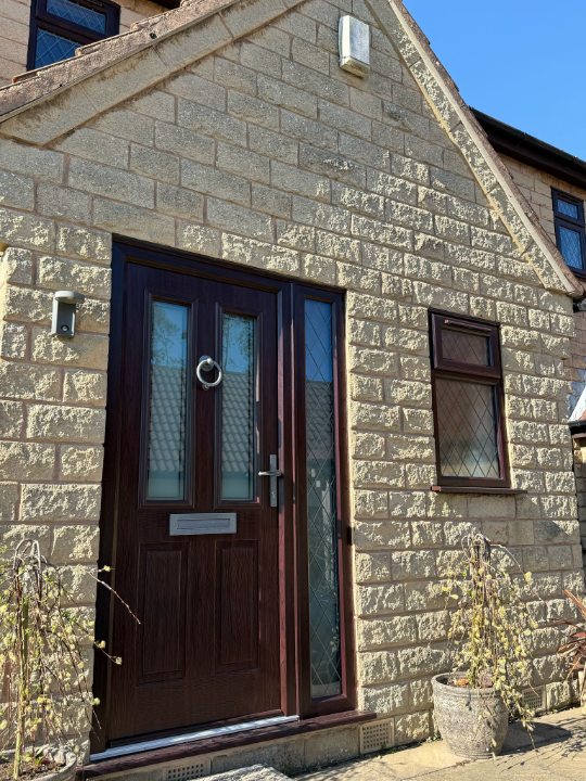 Discover this charming modern home with light-colored stone brick facade, dark wood front door, and lattice-pane windows on a sunny day. Features a classic silver knocker, outdoor light, and decorative potted plants. Perfect for real estate, architecture, and home design inspiration.