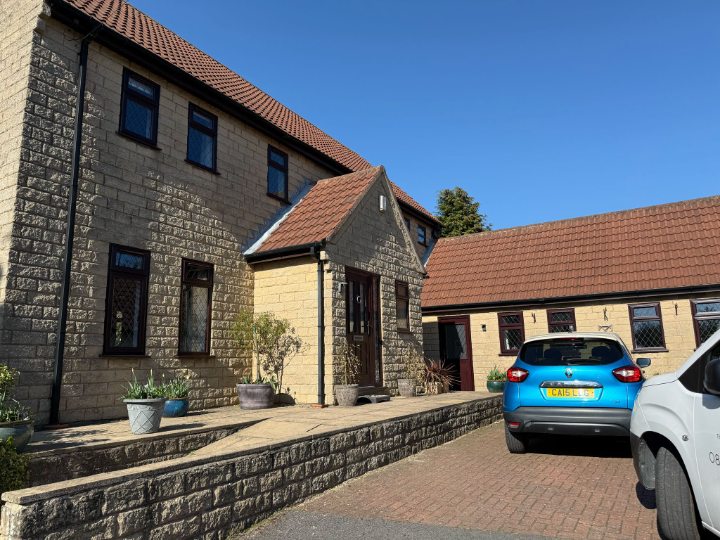 Exterior view of a modern suburban house with light stone facade, red tiled roof, and a blue Renault Captur parked on the brick driveway under a clear blue sky. Features landscaped porch area and retaining wall.